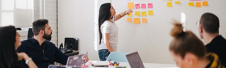 Person steht vor einer Tafel und zeigt auf Klebezettel, Personen sitzen an einem Tisch